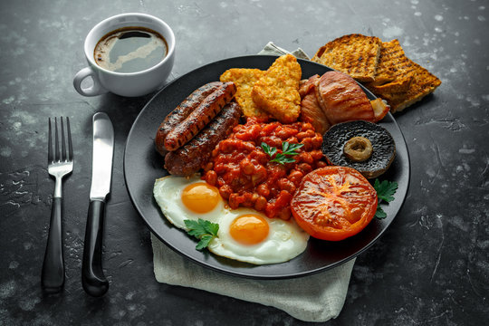 Full English breakfast with bacon, sausage, fried egg, baked beans, hash browns and mushrooms in black plate. cup coffee.