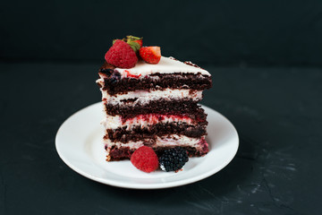 Cut slice of cream cake. On black background and a white saucer, with strawberry raspberries. Dietary white from cream, with chocolate layers of berries.