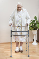 Bardzo stara kobieta w białym szlafroku i koszuli nocnej idzie przy pomocy balkonika rehabilitacyjnego 