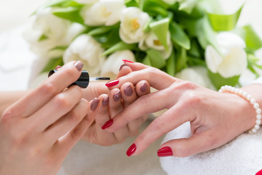 Close-up of the hands of a skilled manicurist, applying elegant red nail polish on the medium length nails of a young woman in a trendy beauty salon