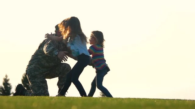 Soldier meet his daughters in the park, side view. Daughters come to their military father in camoubackgrounde and hug him.