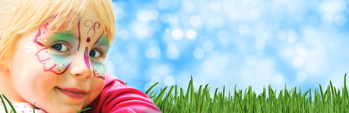 glückliches geschminktes Kind in einem Freizeitpark liegt auf Rasen mit sommerlichem Hintergrund