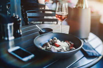 Tilt-shift view: setout in street cafe: clay plate with delicious salad with beef stew and cherry tomatoes, gravy-boat with sauce, glass and bottle of exquisite wine, smartphone, cutlery in soft cover