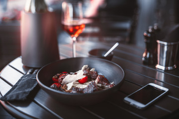 True tilt-shift shooting of table in street restaurant served with clay plate with delicious meat salad with cherry tomatoes, wine bottle, and glass; smartphone and cutlery in soft case near plate