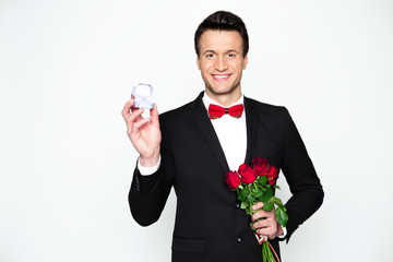 Handsome confident modern elegant man in a official black suit and a red bow tie is holding a box with an engagement ring and beautiful red roses.