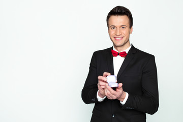 Handsome smiling modern elegant man in a official black suit and a red bow tie is holding a box with an engagement ring and shows in camera.