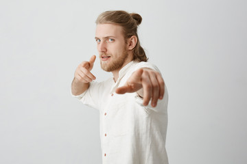 Confident macho man with blond hair and bun, pointing at camera with index fingers, standing half-turned and expressing positive intentions over gray background. Handsome guy acts as if he holding bow