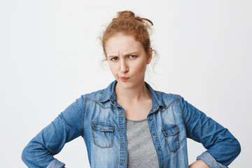 Indoor shot of suspicious frowning redhead european girl squinting and folding lips with hands on waist, expressing disbelief, standing against gray background. I feel like you are hiding something
