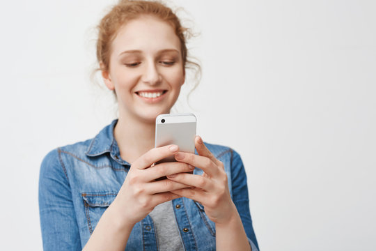 Portrait of positive charming redhead girl with bun hairstyle smiling sensually while holding smartphone and texting, standing against gray background. Barrista plays game while there are no customers