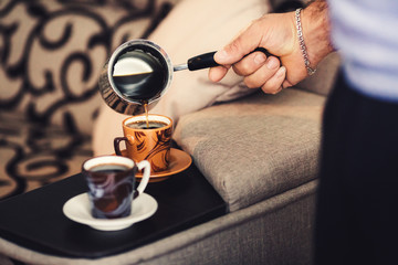 A man's hand pours coffee into a cup on the bedside of the bed. Side view.