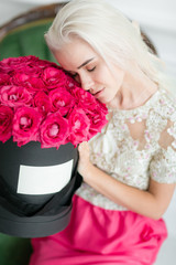 Young pretty girl with cute face and long blond hair. Woman sits and holding black box with pink roses. Luxury interior