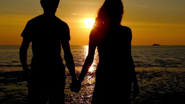 Young couple holding hands walk towards the shore on the beach at sunset in the island of Koh Phangan, Thailand. Valentine Day celebration. Romantic scene, honeymoon love concept