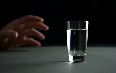 hand reaches for a glass of vodka or alcohol drink, alcoholism and alcohol abuse concept, defocused,.selective focus, close up, gray table, dark background
