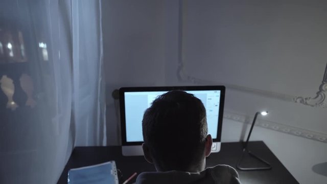 Young author in gray hoodie struggling with screenplay writing on computer at night in old style room, dolly shot