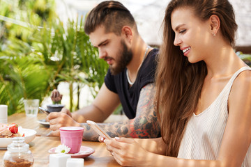 Addicted young female and her bearded handsome male friend send message on smart phone or blog in social networks while sit in outdoor cafe with fresh beverage, connected to wireless internet