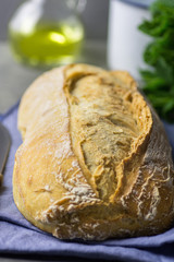 Freshly Baked Hand Crafted Rustic Bread Loaf on Blue  Linen Towel on Dark Kitchen Table Knife Green Herbs Bottle of Olive Oil. Natural Light Authentic Style. Lifestyle Photo. Mediterranean Cuisine