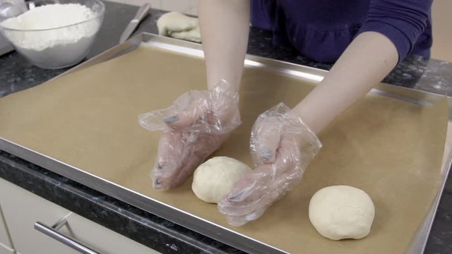 Female baker is putting future buns to the baking pan at the kitchen. The equipment is covered with special brown paper, lady is using her hands in white gloves to cut dought by knife. The glass bowl
