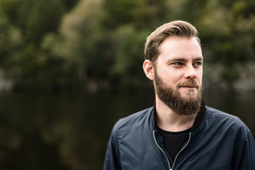 One bearded man wearing a blue jacket, standing in front of a lake and trees on a moody day.