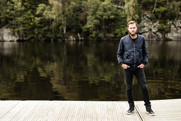 One bearded man wearing a blue jacket, standing in front of a lake and trees on a moody day.