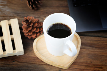 Hot black coffee in white glass, placed on a heart-shaped tray, made of wood and has a corner of a laptop, pine cone is placed around the glass with a romantic color scheme.