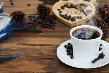 Hot black coffee in white glass placed on wooden floor and feather with heart shaped tray in background.