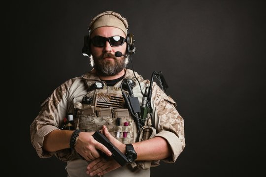 Brutal man in the military desert uniform and body armor stands and holds a gun on a black background in the Studio. The bearded player in the airsoft safety glasses or goggles and active headphones