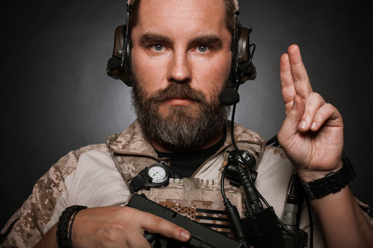 Close-up portrait of brutal bearded man in military desert uniform and body armor who holds his gun on black background. Studio photography of a player in airsoft