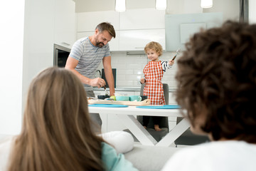 Portrait of father and son cooking together in modern kitchen with mother and sister watching them