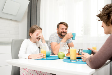 Obraz na płótnie Canvas Portrait of carefree young family laughing happily while enjoying breakfast at home sitting at dinner table in cozy kitchen in morning, drinking orange juice
