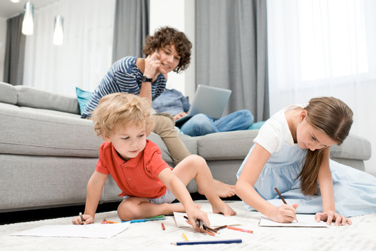 Portrait of two children drawing pictures sitting on carpet in living room with mom watching them from sofa
