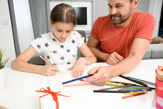 Portrait of bearded father helping little girl drawing handmade card for mothers day at home in modern living room