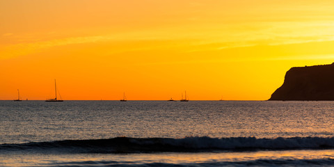 Sail boats anchored near a cliff and a beach at sunset with a beautiful orange sky