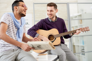 Portrait of two carefree young men having fun in office playing guitar and singing songs sitting on desk