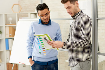 Portrait of two young men, one of them Middle-Eastern, discussing color palette for pamphlet design standing in modern office, copy space