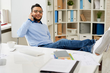 Full length portrait of modern Middle-Eastern entrepreneur relaxing in office speaking by phone with legs on desk and smiling confidently at camera, copy space