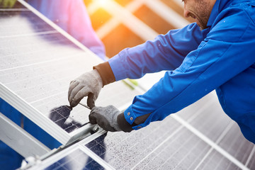 Smiling male technician in blue suit installing photovoltaic blue solar modules with screw. Man...
