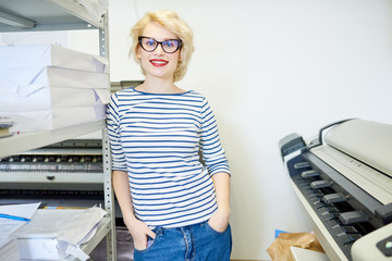 Portrait of blonde young woman standing by plotter machine and smiling at camera while posing in...