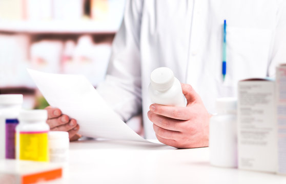 Pharmacist reading prescription with medicine and pill bottle in hand in pharmacy. Medical professional in drugstore counter. Clerk or druggist in drug store. Pharmaceutical retail business concept.