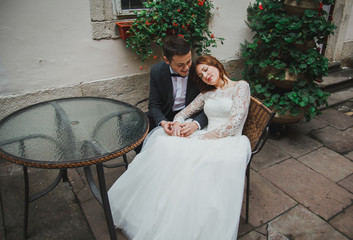 Wedding couple sits in old city vintage street cafe. Stone walls, flowers and glass table in...