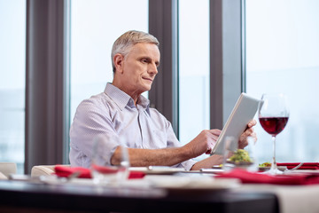Useful tablet. Nice pensive mature man carrying tablet while looking at the screen and drinking wine