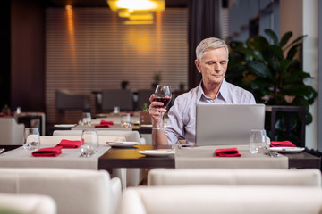 Fototapeta na wymiar Pleasant evening. Concentrated earnest mature man holding wine while staring at the laptop and resting at the restaurant