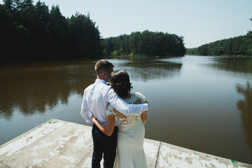 Wedding couple hugs and embraces on the wooden pier. They look at beautiful view over the lake. Sunny summer day for groom and bride in elegant lace dress.