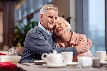 Strong shoulder. Adorable nice mature couple hugging while smiling and sitting at the table