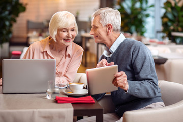 Look at this. Merry gay mature couple sitting at restaurant and smiling while man pointing at tablet