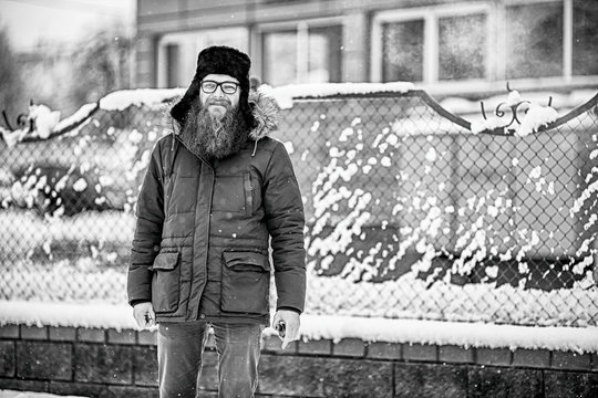 Vape bearded man in real life. Portrait of young guy with large beard in glasses and a cap standing with an electronic cigarette opposite the fence in the winter. Black and white.