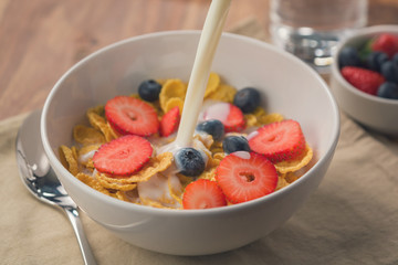 pour milk into corn flakes with berries in white bowl for breakfast on table
