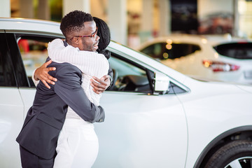 Beautiful young african couple is smiling, looking at camera and hugging while buying car in motor show - obrazy, fototapety, plakaty