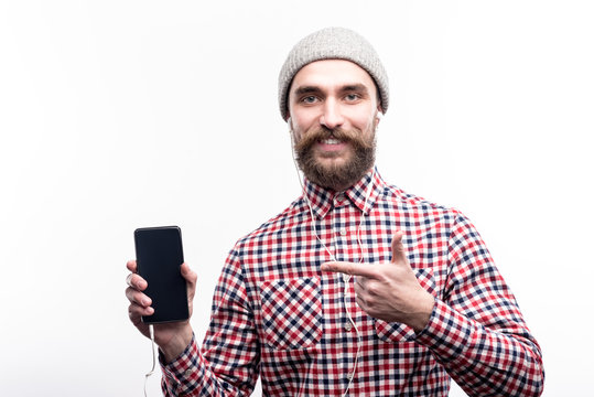 Upbeat listener. Joyful young man in a grey beanie listening to the music in headphones and pointing at a phone in his hand with a finger