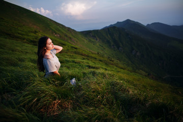 Naklejka na ściany i meble A girl sits on the edge of the cliff and looking at the sun valley and mountains. Woman sitting on mountain top and contemplating the sunset. Young woman in long white dress