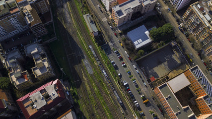 Aerial perpendicular view of the tracks of a train station passing by a road where there are cars parked. A few bushes of grass grows between the rails. There is nobody.